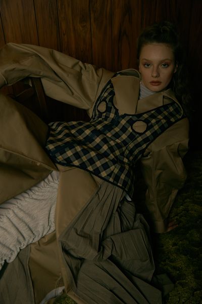 A model sitting on the floor leaning back in a dark room, wearing an olive green coat under a checkered-green top.