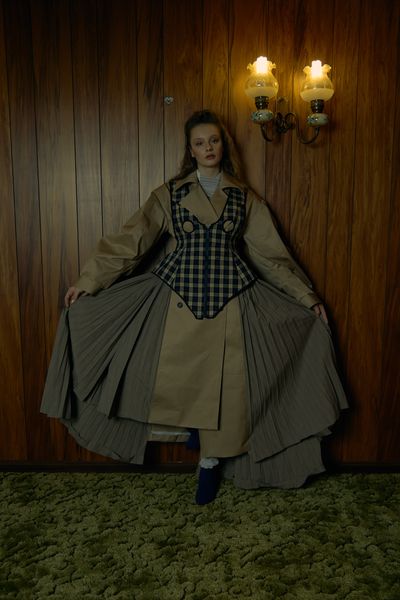 A model standing against a wood panelled wall with an antique light fixture, wearing an olive green coat under a checkered-green top. The model is pulling the bottom half of the coat outwards revealing the different types of fabric it is comprised of.