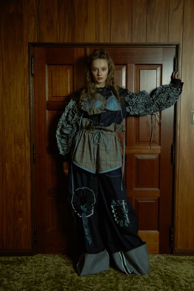 A model standing against a wooden door set into a wood panelled wall, with their arm out leaning against the door frame while wearing a dark long sleeve denim outfit beneath a light blue top.