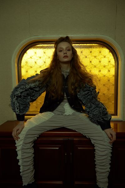 A model sitting on a cabinet in front of a yellow, old-style frosted glass window. The model is wearing a white & beige top and matching pants with a black & blue coat made from different types of material, sitting with their legs spread wide looking directly at the camera.