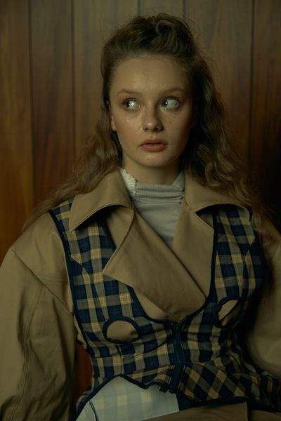 A model standing in front of a wooden wall, facing the camera with their eyes looking to the side. The model is wearing a beige trenchcoat beneath a checkered vest.