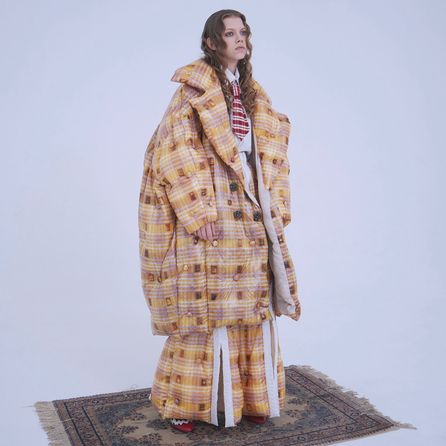 A model standing in a completely white room wearing an oversized yellow & brown tartan jacket and matching oversized pants. The model is looking off in the distance while standing on a faded persian rug.