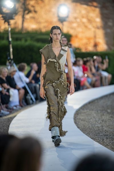 A model walking along a circular catwalk wearing an outfit consisting of parts of an olive green jacket and lighter components with distressed seams where each component connects.