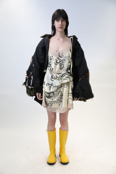 A model standing against a white backdrop wearing a gold silken nightwear with black swirls beneath a large jacket open to show the black lining and outer gold & black pattern. The jacket has multiple distressed seams where different fabrics that comprise the jacket meet. The model is also wearing vivid yellow gumboots.