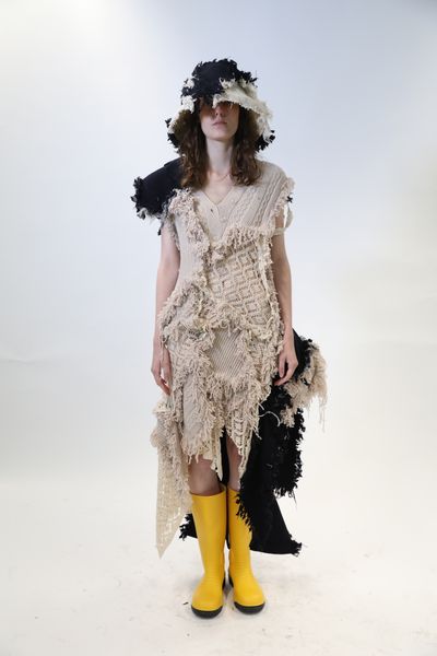 A model standing against a white backdrop wearing a full-length cream-coloured outfit comprised of multiple knitwear fabrics joined together by rough, distressed seams. The shoulder and one side also incorporate contrasting black segments. The model is also wearing a matching distressed cream & black bucket hat that extends down near their shoulders. The model is also wearing vivid yellow gumboots.