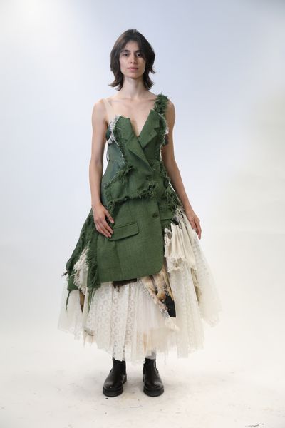 A model standing against a white backdrop wearing an outfit primarily comprised of pieces from a green jacket atop a large, frilly dress incorporating multiple light coloured fabrics.