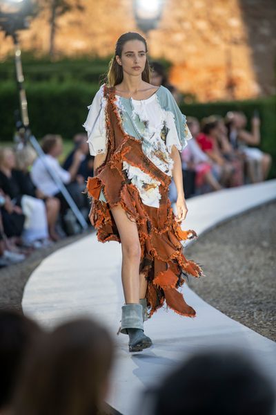 A model walking along a circular catwalk wearing an outfit comprised of multiple pieces of white and light-blue fabric surrounding a large orange segment running diagonally from the shoulder to the opposite side's ankle.