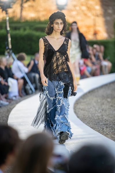 A model walking along a circular catwalk wearing a lacy partially-transparent top comprised of multiple pieces of black fabric, and jeans comprised of multiple pieces of blue denim, joined with rough, distressed seams.