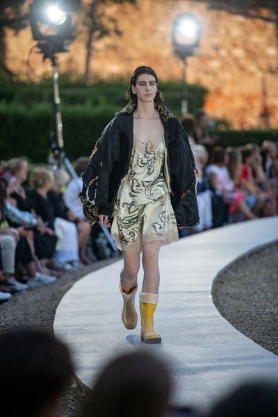 A model walking along a circular catwalk wearing a gold silken nightwear with black swirls beneath a large jacket open to show the black lining and outer gold & black pattern. The jacket has multiple distressed seams where different fabrics that comprise the jacket meet.