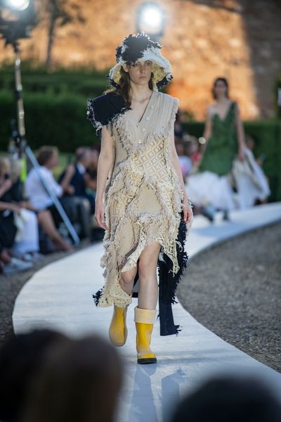 A model walking along a circular catwalk wearing a full-length cream-coloured outfit comprised of multiple knitwear fabrics joined together by rough, distressed seams. The shoulder and one side also incorporate contrasting black segments. The model is also wearing a matching distressed cream & black bucket hat that extends down near their shoulders.