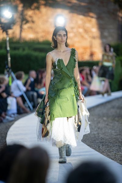 A model walking along a circular catwalk wearing an outfit primarily comprised of pieces from a green jacket atop a large, frilly dress incorporating multiple light coloured fabrics.