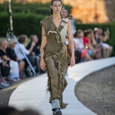 A model walking along a circular catwalk wearing an outfit consisting of parts of an olive green jacket and lighter components with distressed seams where each component connects.